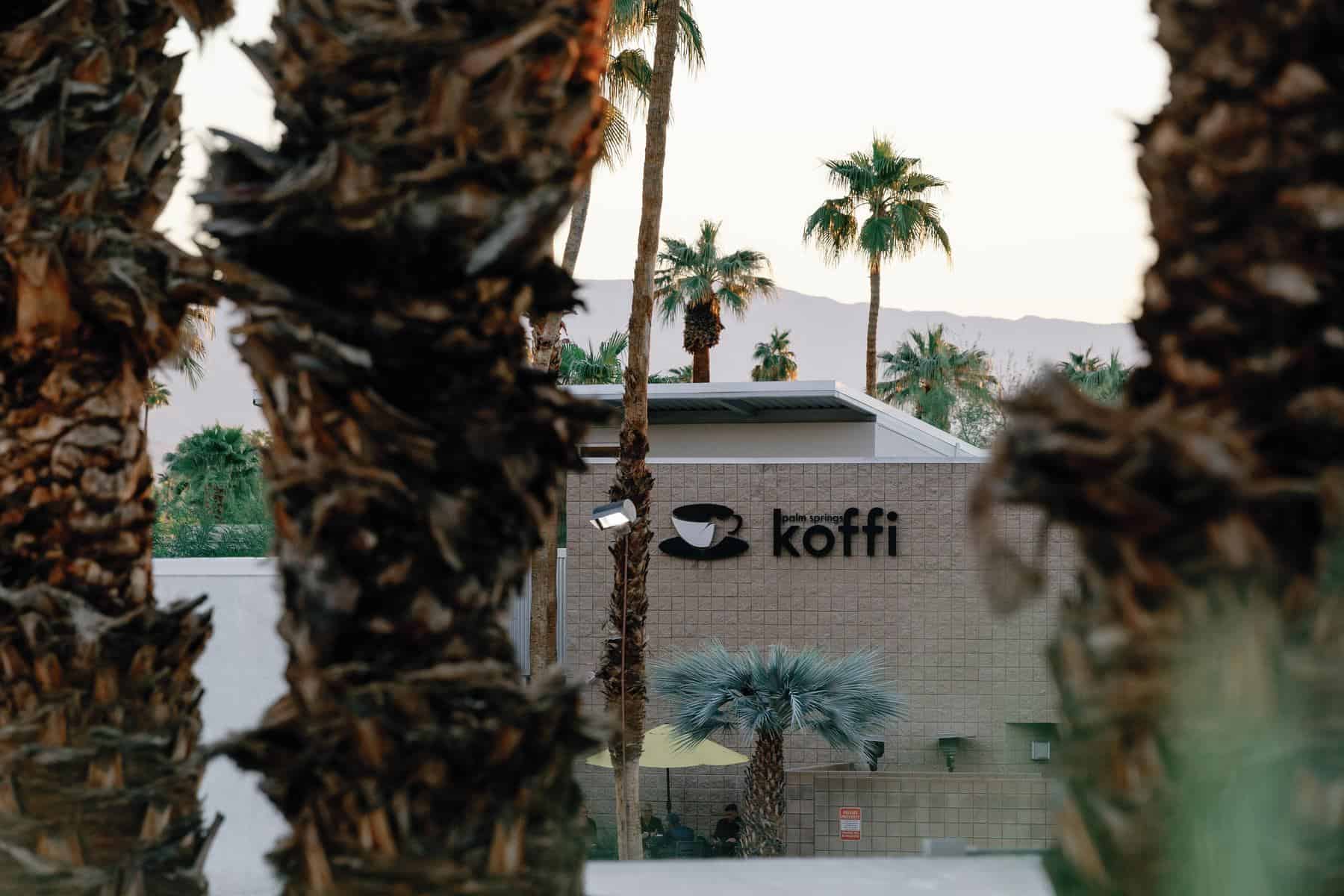 Koffi South in Palm Springs, with umbrella patio seating, seen through palm trees with mountains behind